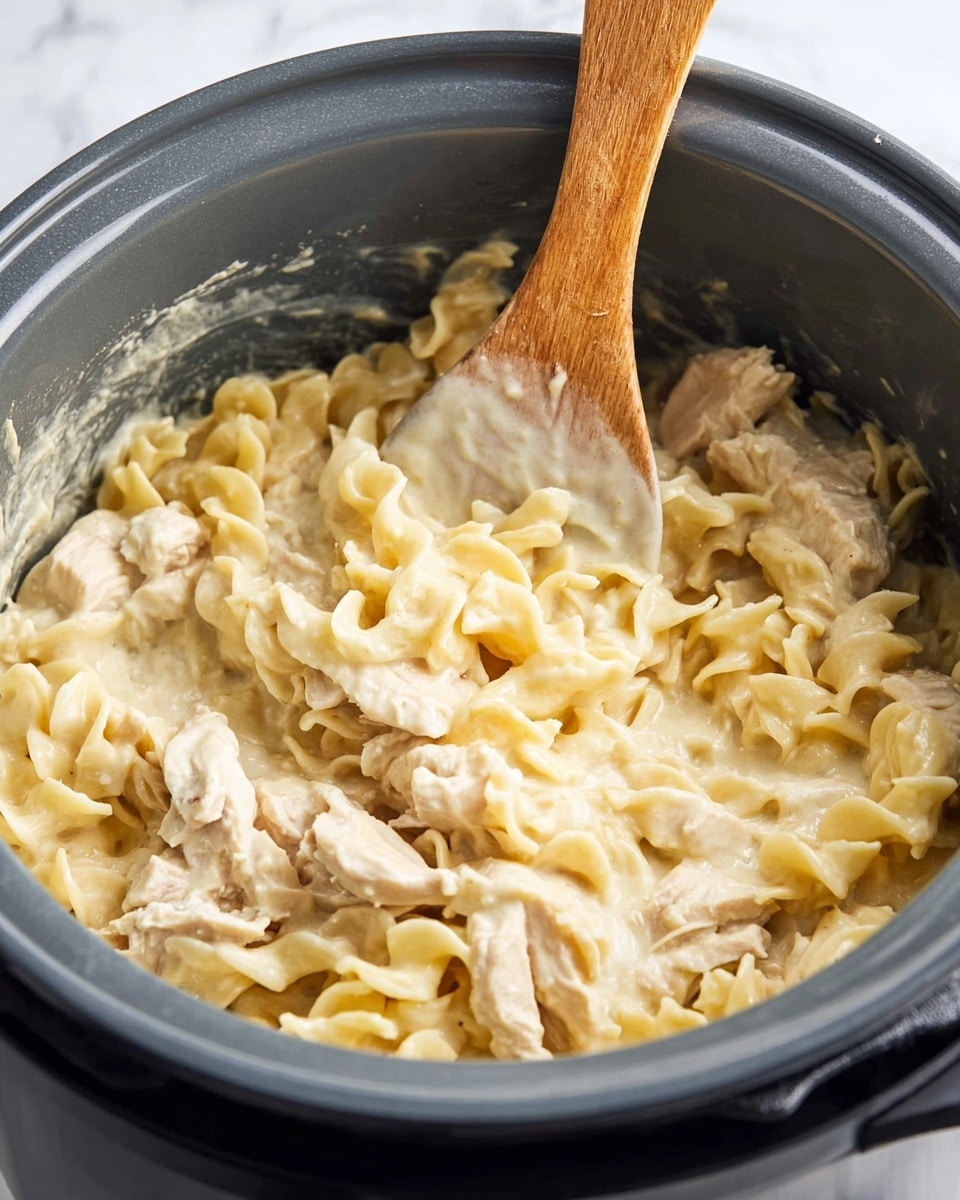 Inside a dark gray pot, there is a creamy pasta dish made of wavy egg noodles covered in a thick, pale yellow sauce. The noodles are mixed with chunks of light-colored cooked chicken. A wooden spatula with some sauce on it rests inside the pot, standing vertically. The background shows a white marbled texture. photo taken with an iphone --ar 4:5 --v 7