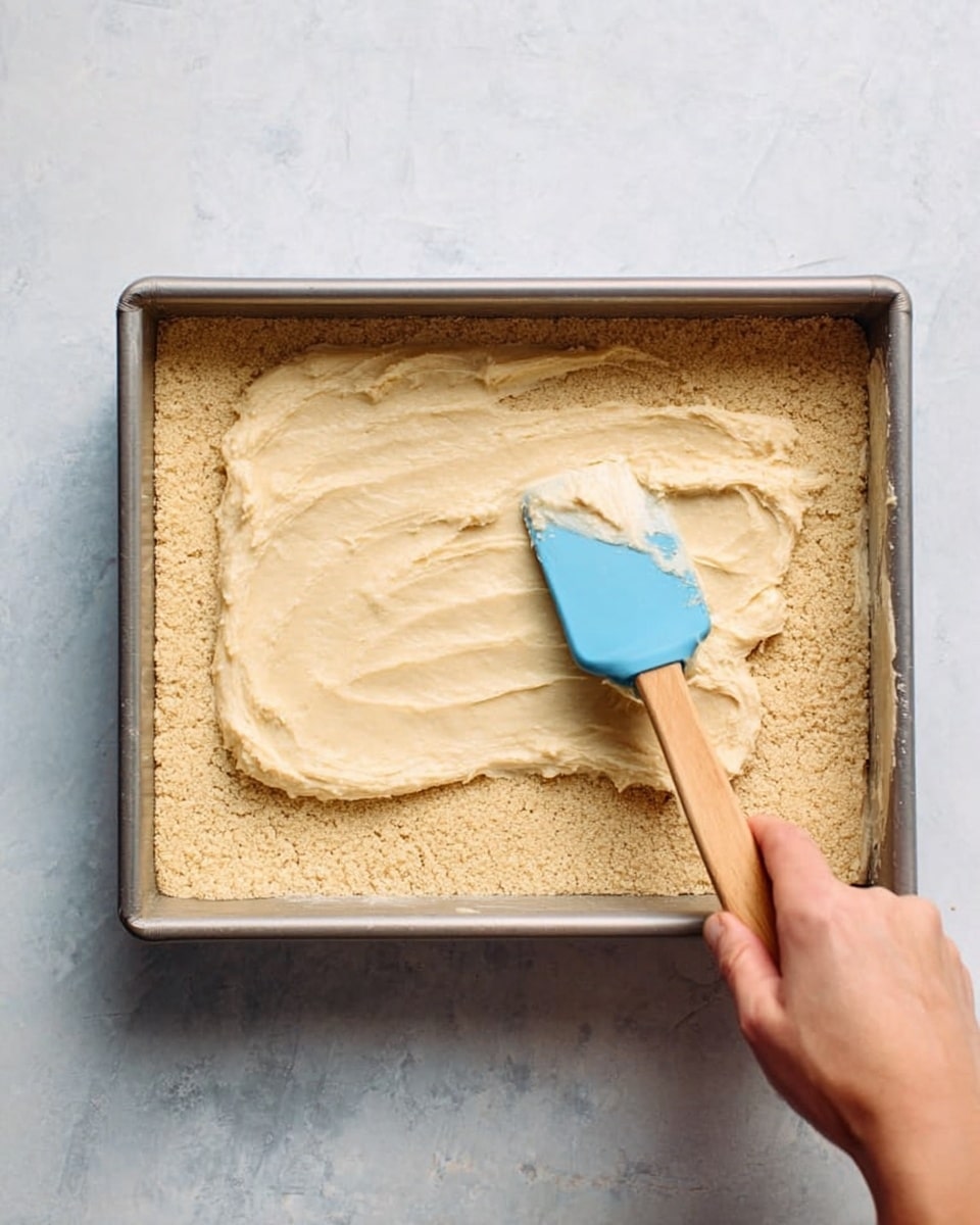 A rectangular metal pan holds a thick, light golden crust layer that fills the bottom completely with a slightly rough texture, resembling a baked dough base. On top, there is a generous dollop of creamy, smooth beige mixture that is being spread with a blue silicone spatula featuring a wooden handle. A woman's hand grips the spatula, partially visible from the right side. The pan rests on a white marbled surface, adding a clean and bright background to the scene photo taken with an iphone --ar 4:5 --v 7