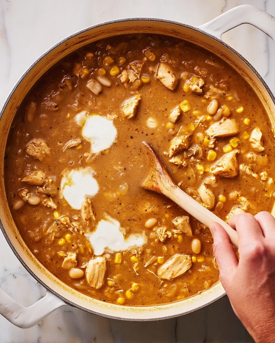 A large white pot filled with a thick, brown stew that has chunks of light brown cooked chicken, small yellow corn kernels, and white beans scattered throughout. There are two white dollops of creamy sauce melting into the stew near the center. A woman's hand is stirring the stew with a wooden spoon, creating a smooth motion in the mixture. The pot sits on a white marbled surface. photo taken with an iphone --ar 4:5 --v 7