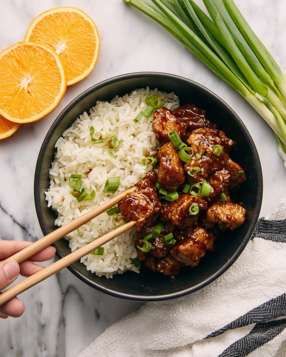 A black bowl holds two layers of food on a white marbled surface. The left half has white cooked rice sprinkled with small slices of green onion. The right half has glossy brown cooked chicken pieces coated in sauce, also topped with green onion pieces. Two light wooden chopsticks held by a woman's hand pick up one piece of the chicken. Next to the bowl are three orange slices on the white marbled surface and a bunch of green onions at the top right. A white cloth with black stripes is partially visible at the bottom right. Photo taken with an iphone --ar 4:5 --v 7