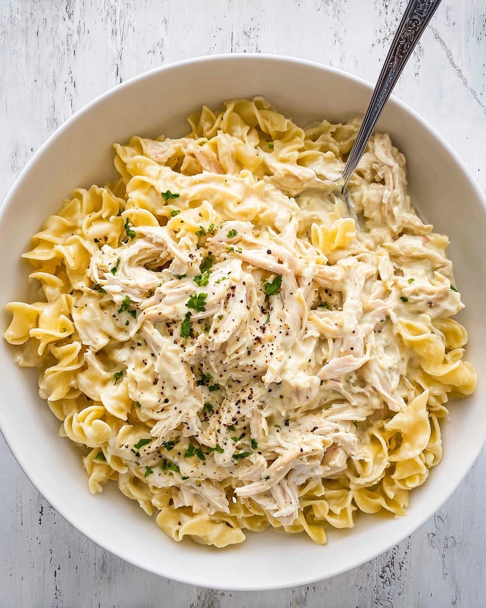 A white bowl filled with creamy chicken pasta sits on a white marbled surface. The dish has three main layers: a base of soft, light yellow twisted egg noodles, a middle layer of shredded pale white chicken pieces mixed with the noodles, and a top layer of creamy white sauce coating everything evenly. Small bits of green herbs are sprinkled on top, adding a touch of color, along with tiny black pepper specks. A silver fork stands upright in the bowl, partly buried in the pasta. photo taken with an iphone --ar 4:5 --v 7