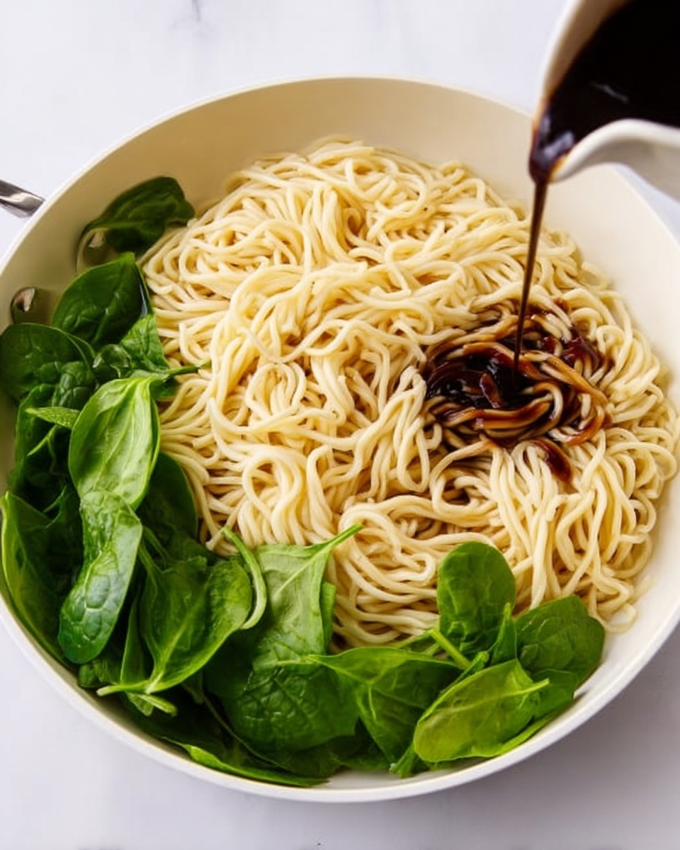 A white bowl with cooked pale yellow noodles piled loosely inside, arranged in the center and spreading outward. Fresh bright green spinach leaves sit on one side of the noodles, adding a vibrant contrast. Dark brown sauce is being poured onto the noodles from above, pooling slightly in the middle and coating some strands. The bowl rests on a clean white marbled surface, creating a fresh and simple background. photo taken with an iphone --ar 4:5 --v 7