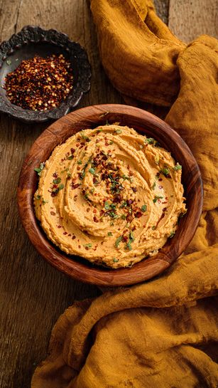 The image shows a round wooden bowl filled with a creamy, light orange spread with a smooth texture and some green herb pieces mixed in. The bowl is placed on a crumpled mustard yellow cloth, which lies on a wooden surface. Next to the bowl, there is a small dark dish containing a mix of spices in red and brown tones. The scene is warm and rustic, focusing on the creamy spread in the bowl. Photo taken with an iphone --ar 4:5 --v 7