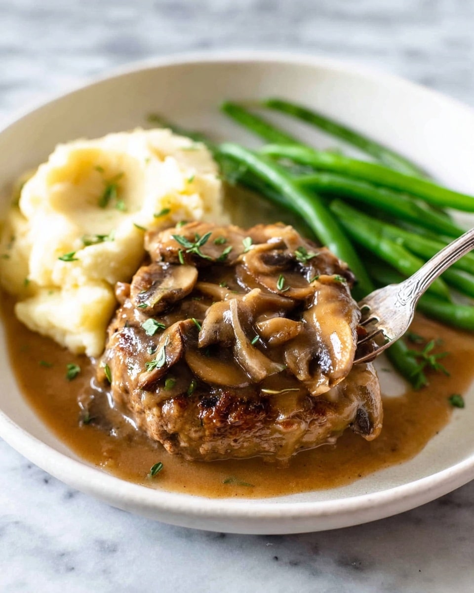 The image shows a white round plate with three main layers of food on a white marbled surface. The first layer is a serving of creamy mashed potatoes with a smooth, pale yellow texture, placed on the left side of the plate. On top of the mashed potatoes is a thick, browned meat patty covered in a rich brown mushroom gravy, with visible slices of cooked mushrooms and small green herbs sprinkled on top. To the right of the meat patty are bright green, glossy cooked green beans, adding a fresh contrast in color and texture. A silver fork is picking up a piece of the meat patty near the front of the plate. Photo taken with an iphone --ar 4:5 --v 7
