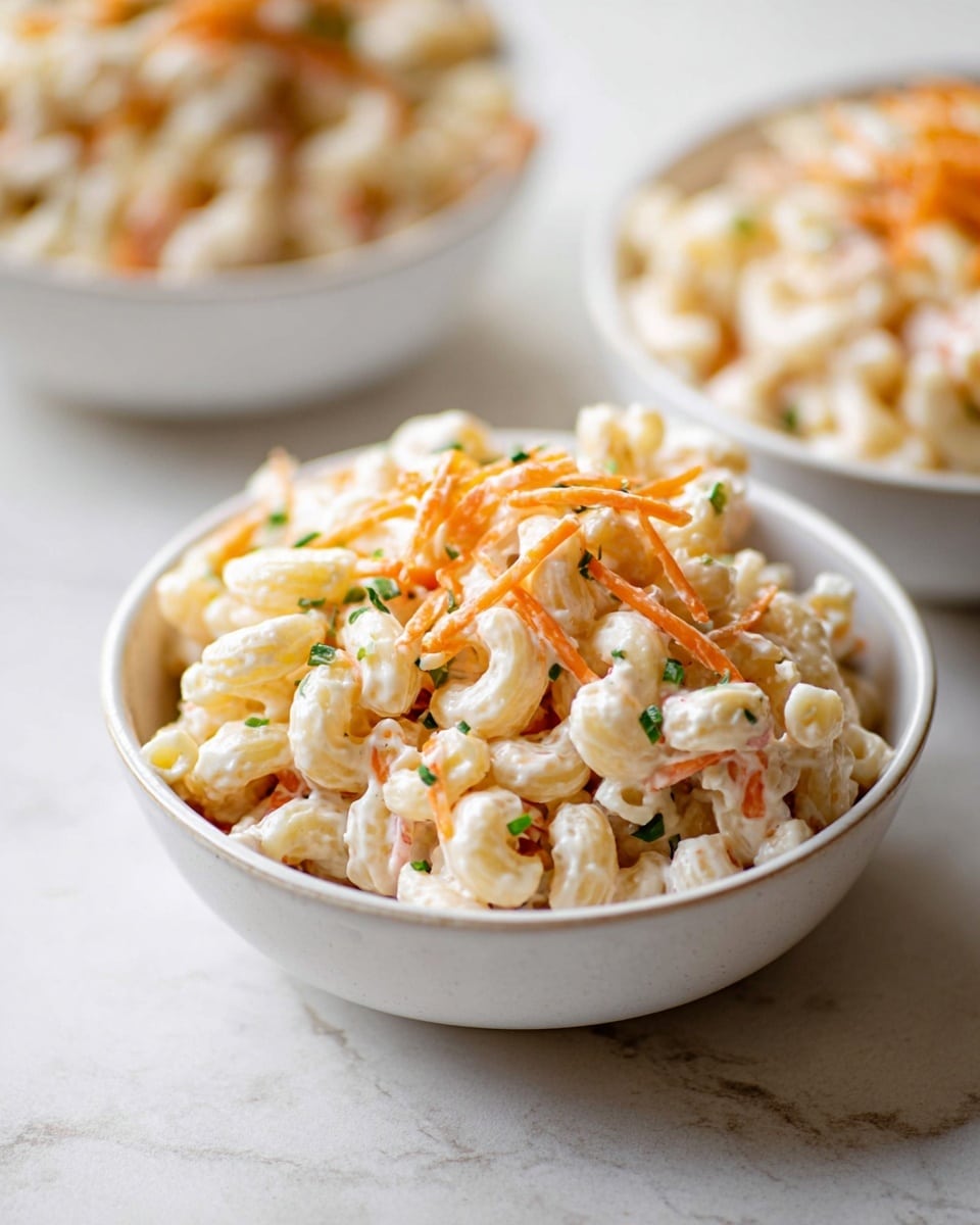 A white bowl filled with creamy macaroni salad made of elbow-shaped pasta in a smooth white dressing. On top, there are thin orange strips of carrot and small green herb pieces sprinkled around. The bowl is placed on a white marbled surface with two more similar white bowls blurred in the background, each also filled with the same macaroni salad. The pasta looks soft, slightly shiny from the dressing, and the overall color is light with pops of orange and green. Photo taken with an iphone --ar 4:5 --v 7