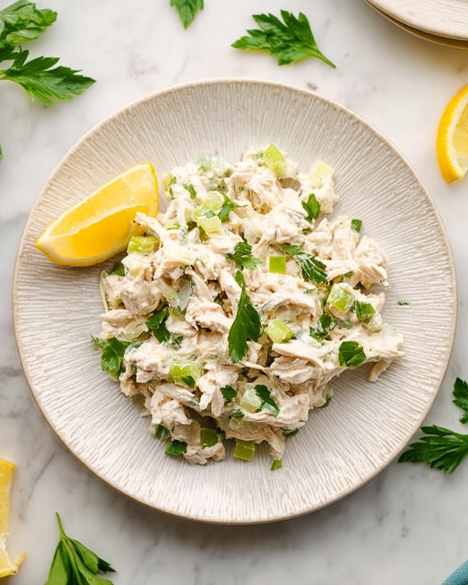 A white plate with a textured rim holds a creamy chicken salad. The dish has pale white shredded chicken pieces mixed with small green celery chunks and fresh green parsley leaves spread evenly throughout. On the left side of the plate, there is a single bright yellow lemon wedge placed next to the salad. The plate sits on a white marbled surface with a few loose parsley leaves scattered around and some lemon wedges in the background. The image is bright and clear, showing the textures of the creamy salad and fresh greens. photo taken with an iphone --ar 4:5 --v 7