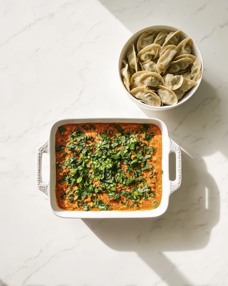 A white square baking dish with handles holds one layer of thick orange sauce, topped with chopped fresh green leafy vegetables spread evenly across the surface. Above and to the right, there is a white bowl filled with many small, half-moon shaped dumplings that are light beige in color, showing a soft texture. The whole setup is placed on a white marbled textured surface, with bright, natural light casting soft shadows. Photo taken with an iphone --ar 4:5 --v 7