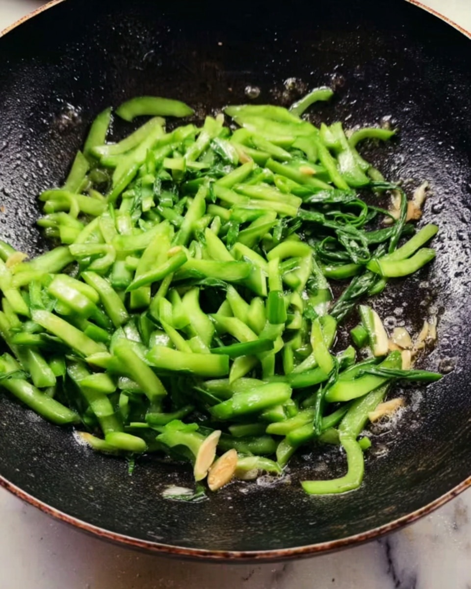 The image shows green sliced vegetables cooking in a black frying pan. The vegetables are bright green with a smooth texture and are mixed with small pieces of light-colored garlic or ginger. The frying pan has a slightly shiny, oily surface with some steam and oil visible. The background is a white marbled surface. photo taken with an iphone --ar 4:5 --v 7