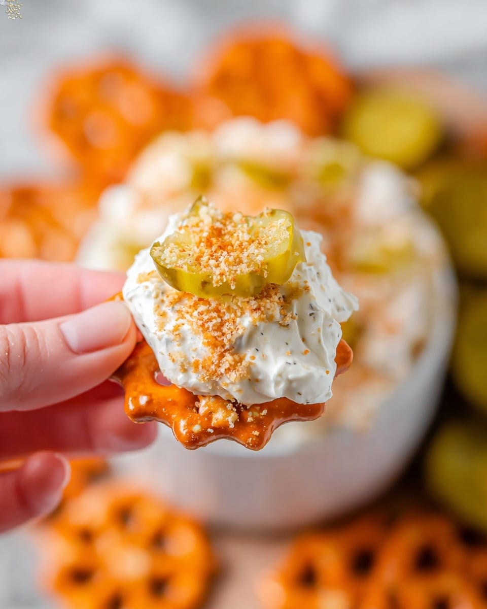 A round white bowl is filled with a creamy white dip topped with a thick layer of golden-brown toasted crumbs. On top of the crumbs, three small bright green pickles are placed closely together at the center. Surrounding the bowl on a wooden board are many orange pretzel snacks, with a few green pickles also visible near the bottom edge. The background surface is a white marbled texture photo taken with an iphone --ar 4:5 --v 7
