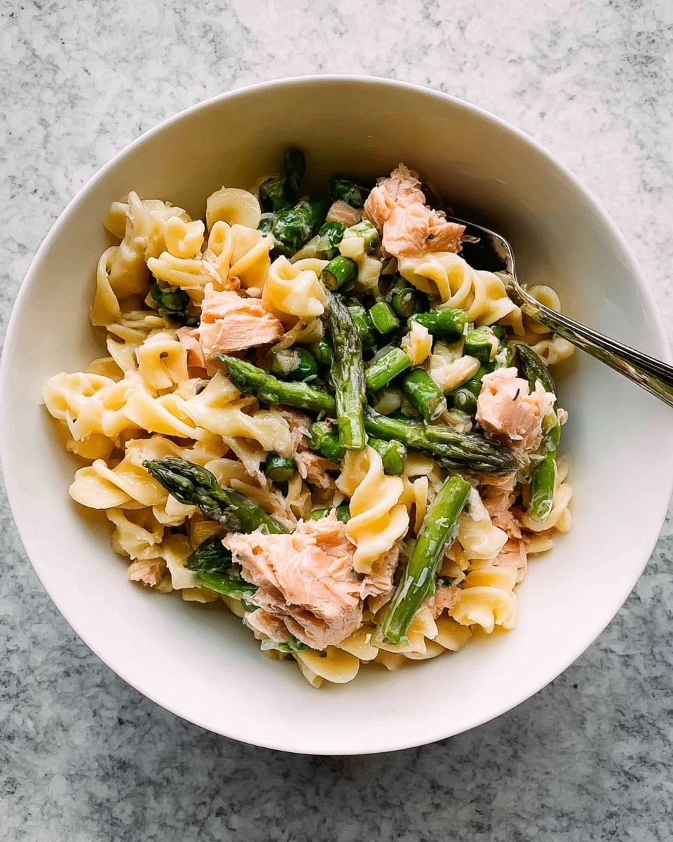 A white bowl filled with three main layers: the first is curly pasta in pale yellow with a smooth creamy coating, next scattered bright green chopped asparagus pieces with a fresh texture, and finally chunks of light pink cooked salmon mixed evenly throughout. A silver fork rests in the bowl among the ingredients, and the bowl sits on a white marbled surface. photo taken with an iphone --ar 4:5 --v 7