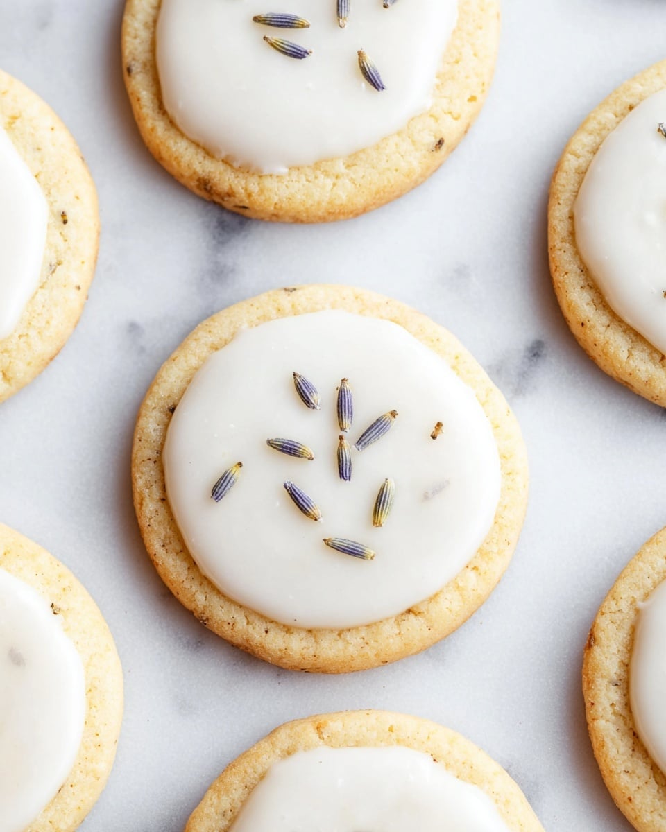 The image shows several round cookies arranged on a white marbled surface, each cookie has a light golden-brown base with a slightly textured surface. On top of each cookie, there is a smooth white icing layer, neatly spread in a circular shape smaller than the cookie base. Each icing layer is decorated with a few small, dried lavender buds placed in the center, adding a subtle touch of color and texture. The cookies are evenly spaced, showing some parts of the white marbled background between them. photo taken with an iphone --ar 4:5 --v 7