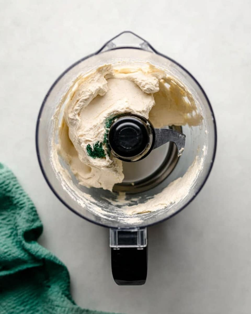 A clear food processor bowl sits on a white marbled surface, filled with a large scoop of smooth, pale cream-colored mixture with small patches of dark green color scattered inside. The blade of the processor is visible beneath the creamy layer, and the black handle is on the right side. A green cloth is partly visible in the upper left corner. The light is soft and natural, giving the cream a slightly shiny texture. photo taken with an iphone --ar 4:5 --v 7