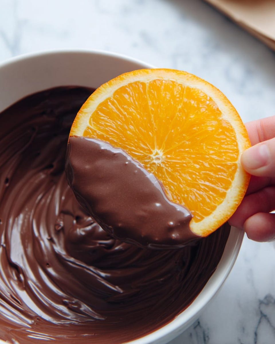A woman's hand is holding an orange slice that is half dipped in thick, smooth dark brown melted chocolate, with glossy texture and gentle folds. The orange slice is bright orange with a translucent, slightly textured surface showing its segments, and the chocolate covers the lower half of the orange slice evenly. The orange slice is above a round white bowl filled with more melted chocolate showing swirls and shiny spots. The scene is on a white marbled surface. Photo taken with an iphone --ar 4:5 --v 7