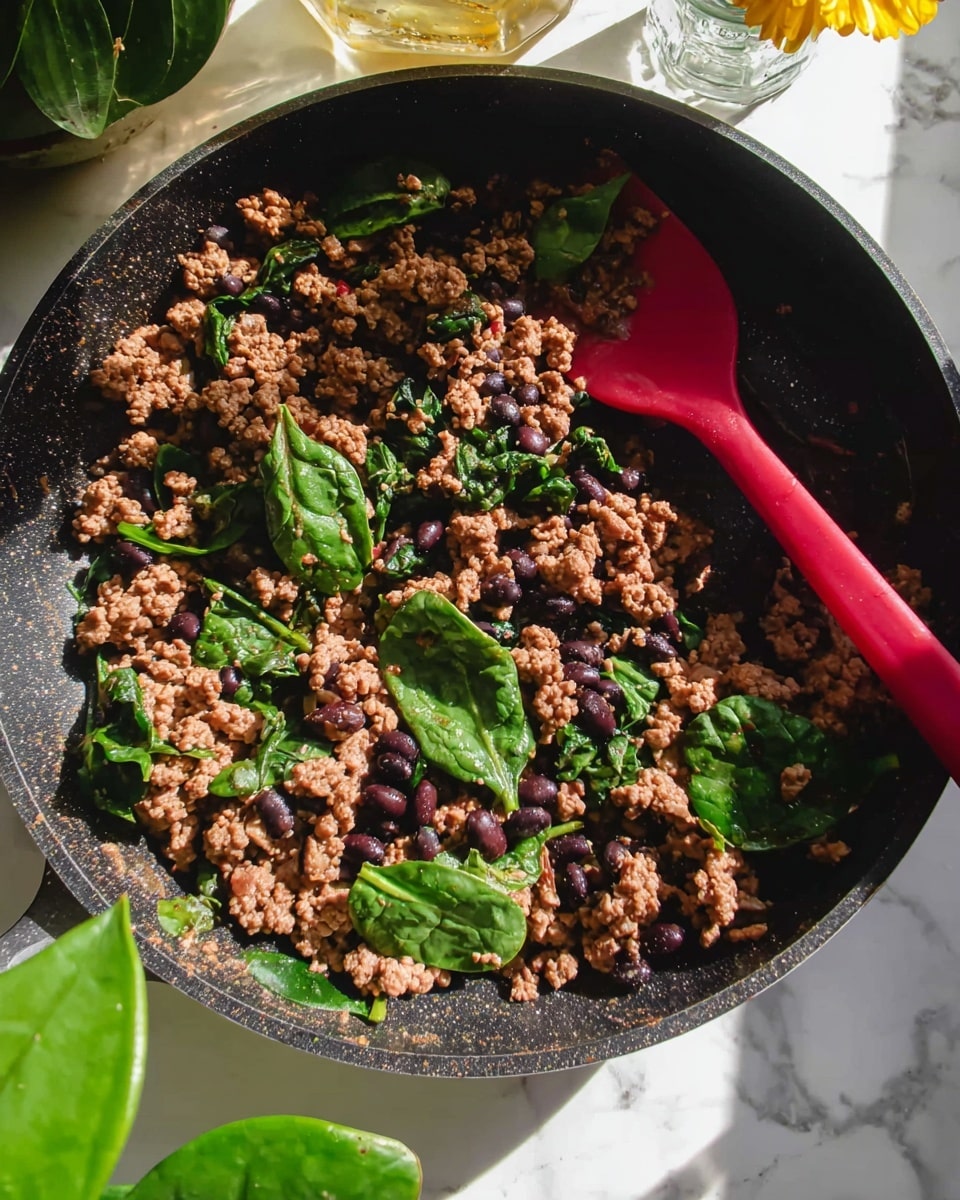 Viral Beef, Sweet Potato and Cottage Cheese Protein Bowls Recipe 4 A close-up of a black pan filled with cooked ground meat mixed with black beans and fresh spinach leaves scattered throughout. The ground meat is brown and crumbly, with black beans adding small, round dark spots inside the mix. Bright green spinach leaves bring color contrast, some whole and some slightly wilted. A red spatula rests inside the pan, partially covered by the meat mixture. The pan sits on a white marbled surface with some sunlight casting soft shadows. In the background, green plant leaves and a partially visible glass jar with a yellow flower add a natural touch. Photo taken with an iphone --ar 4:5 --v 7