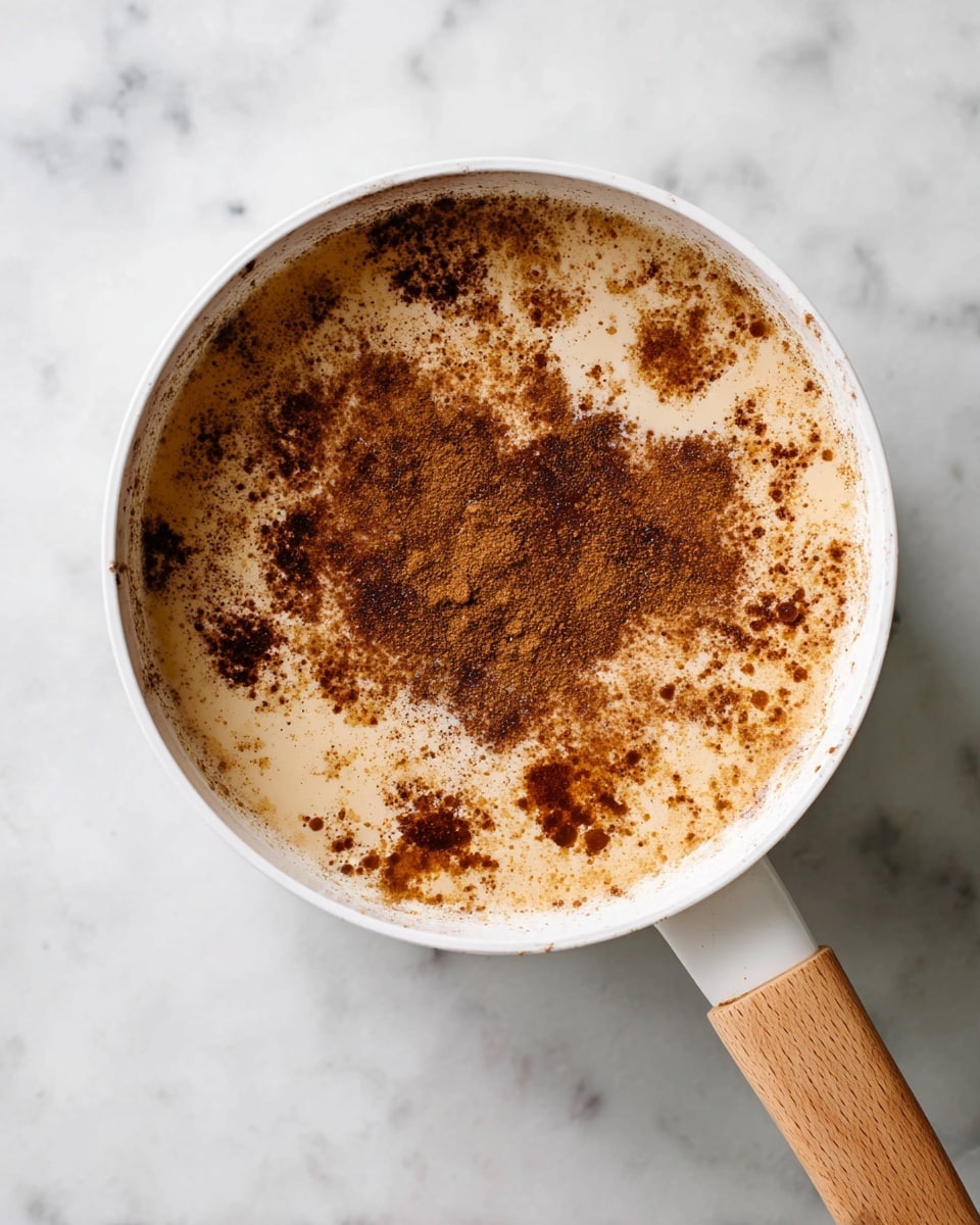 A white pot with a wooden handle sits on a white marbled surface, filled with creamy beige liquid. The top layer is sprinkled unevenly with a generous amount of dark brown cinnamon powder, creating a speckled and textured look with some thicker clumps of cinnamon near the center and lighter dustings around the edges. The mixture appears warm and smooth under the spice cover. photo taken with an iphone --ar 4:5 --v 7