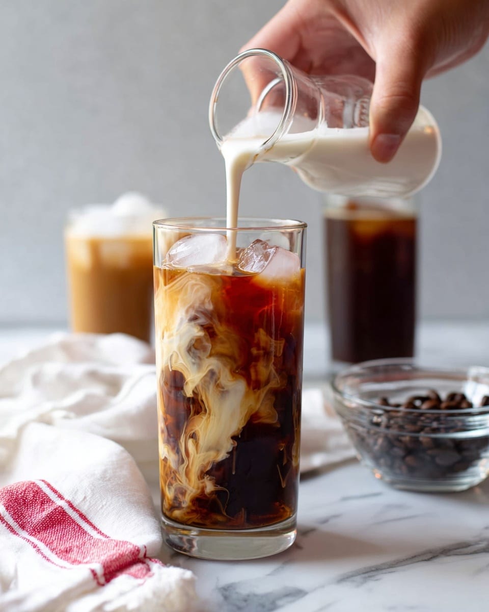 A tall clear glass filled with dark iced coffee containing ice cubes has creamy milk being poured from a small glass jug held by a woman's hand above it. The milk creates swirling white patterns mixing with the coffee, showing layers of dark brown, light brown, and creamy white colors. In the background, there are two blurred glasses also with iced coffee and a small transparent bowl with dark brown coffee beans placed on a white marbled surface. A white cloth with a red stripe is casually draped near the glass. Photo taken with an iphone --ar 4:5 --v 7