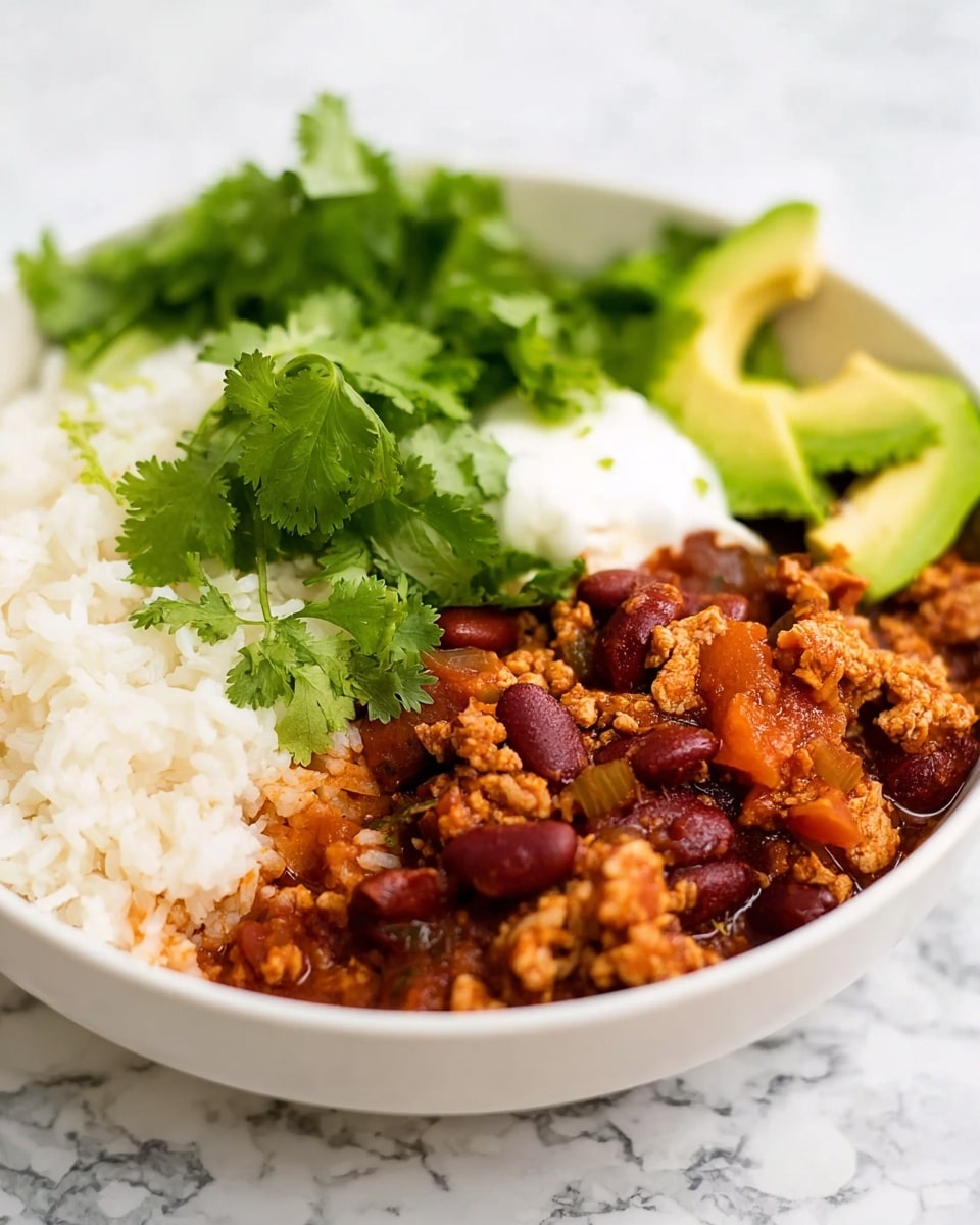A white bowl filled with three main layers is placed on a white marbled surface. On one side, there is a fluffy white rice layer with a light, grainy texture. Next to it is a thick layer of cooked red kidney beans mixed with minced meat pieces in a rich reddish-brown sauce that looks a bit chunky with bits of onions and tomatoes. On top of this layer, there are fresh bright green cilantro leaves and a small dollop of smooth white sour cream, alongside thin slices of green avocado. The bowl's colors stand out nicely against the clean white marbled background, and blurred green herbs appear faintly in the background. Photo taken with an iphone --ar 4:5 --v 7