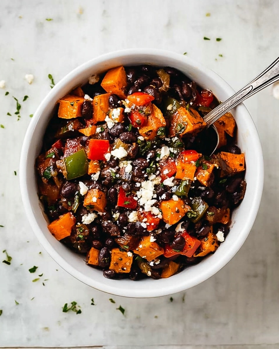 A white bowl filled with a mix of dark black beans, orange sweet potato chunks, and small pieces of red and green bell peppers. There are also bits of green herbs scattered throughout the dish, along with small white cheese crumbs sprinkled on top. A silver spoon rests inside the bowl, and the bowl is placed on a white marbled textured surface with small bits of green herbs and cheese crumbs around. photo taken with an iphone --ar 4:5 --v 7