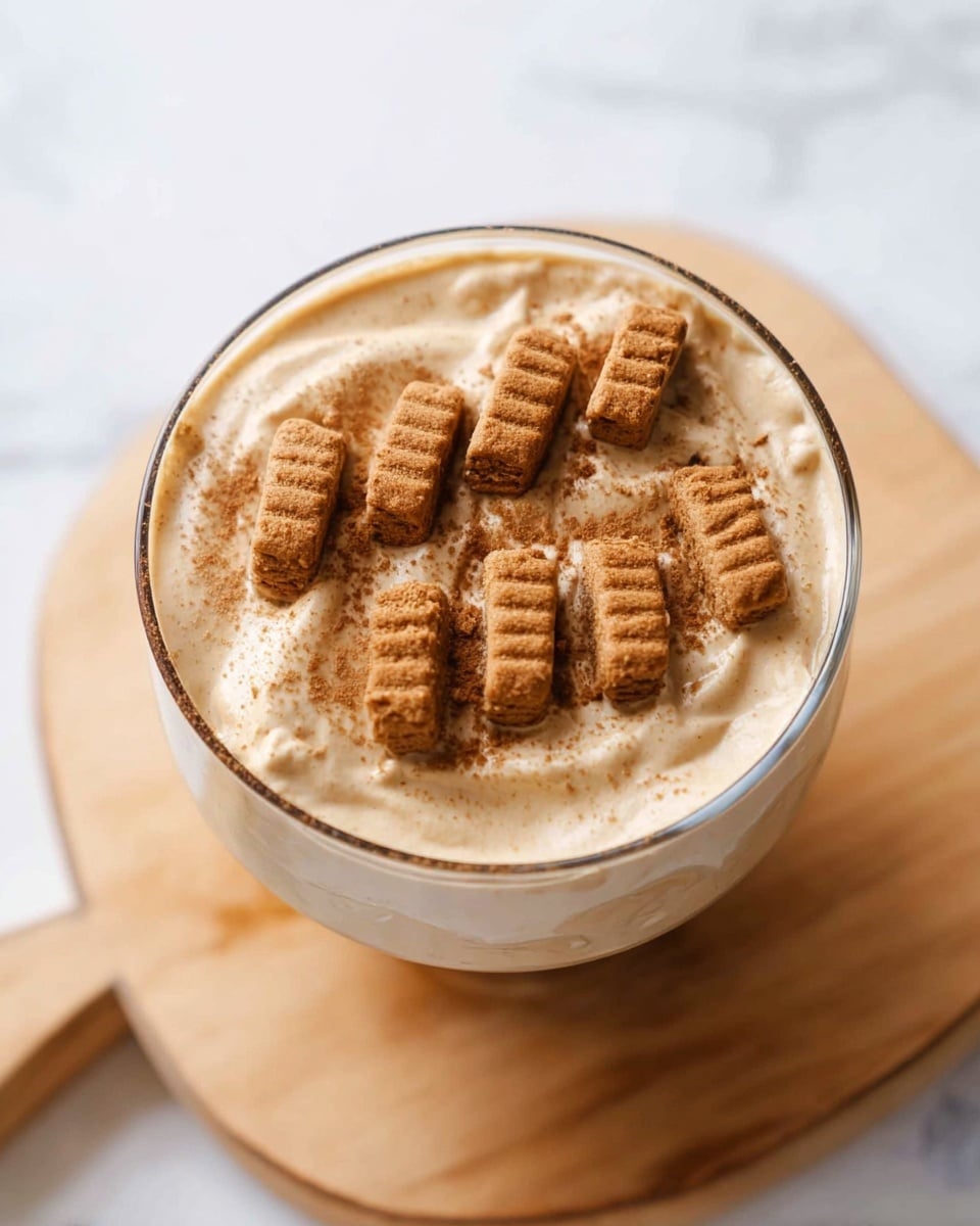 A clear glass bowl filled with a thick, creamy beige dessert topped with eleven small, light brown rectangular cookies placed in two diagonal rows across the surface; the bowl is placed on a light wood cutting board with rounded edges, and the background is a white marbled texture. photo taken with an iphone --ar 4:5 --v 7
