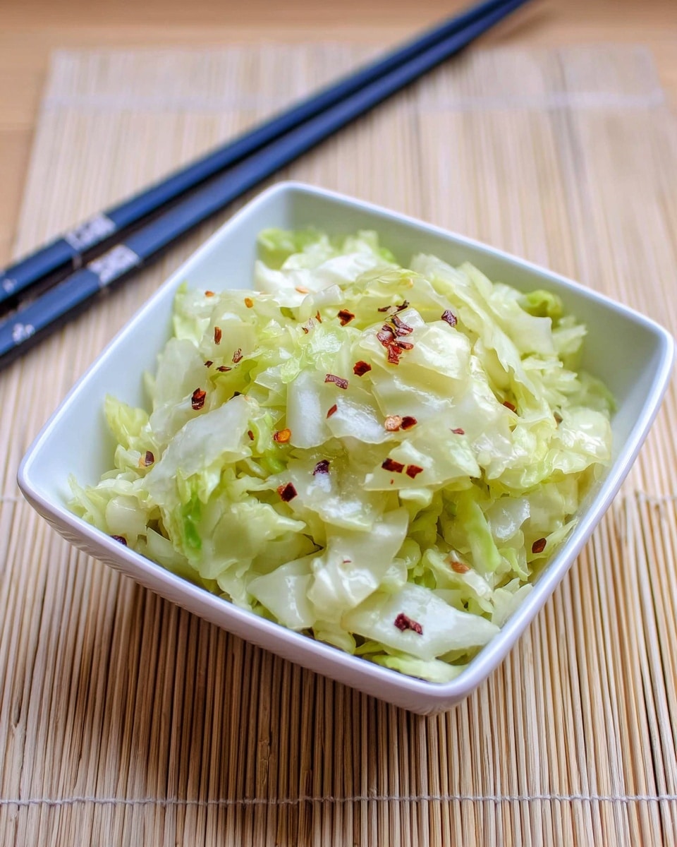 A white square bowl filled with shredded pale green and white cabbage, lightly cooked or steamed, with small red chili flakes sprinkled on top. The bowl rests on a light tan bamboo mat, and a pair of dark blue chopsticks lies next to the bowl. The setting is bright and simple, with the focus on the fresh and slightly soft texture of the cabbage. Photo taken with an iphone --ar 4:5 --v 7