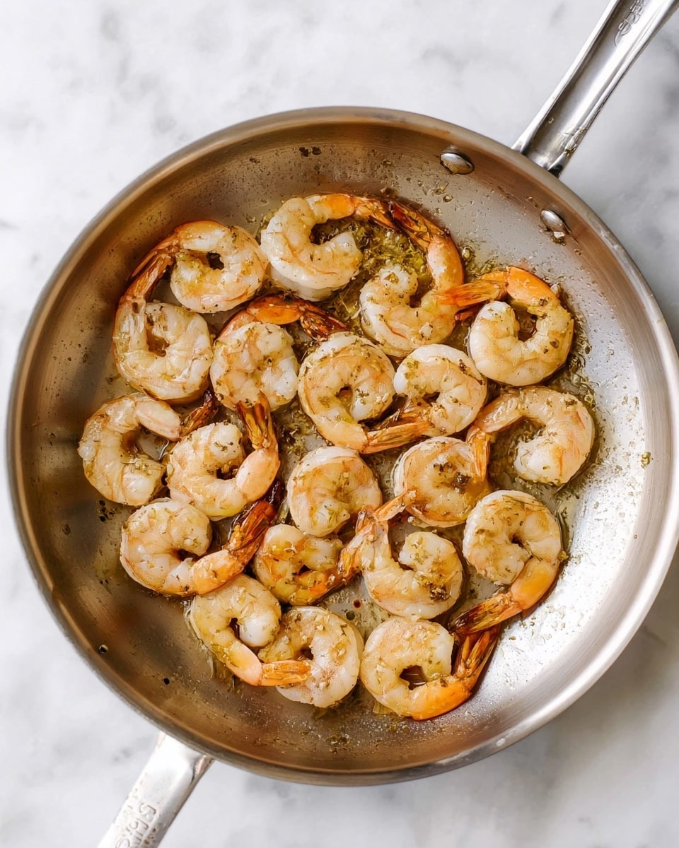 A silver frying pan filled with a single layer of light pink cooked shrimp, some with darker orange tails, arranged in a loose circular pattern. The shrimp have a slight shine and hints of browned spots, showing they are cooked. The pan sits on a white marbled surface, creating a clean and bright background. Photo taken with an iphone --ar 4:5 --v 7