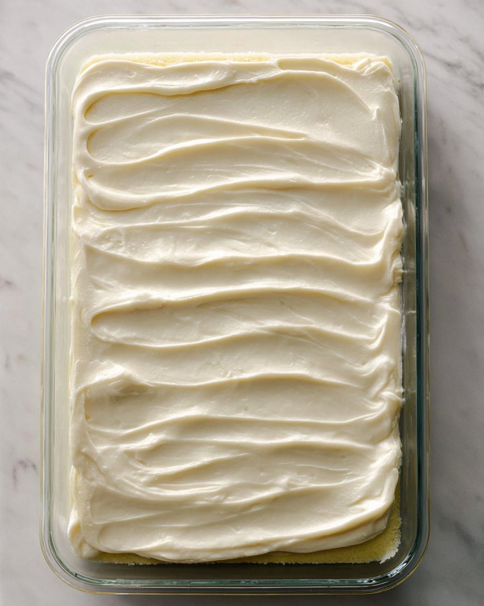 A rectangular glass dish holds a single flat cake layer covered with a thick, smooth layer of creamy white frosting. The frosting is spread evenly with soft, wavy lines running horizontally across the entire surface. The edges show a hint of the pale yellow cake underneath. The dish is placed on a white marbled surface. photo taken with an iphone --ar 4:5 --v 7