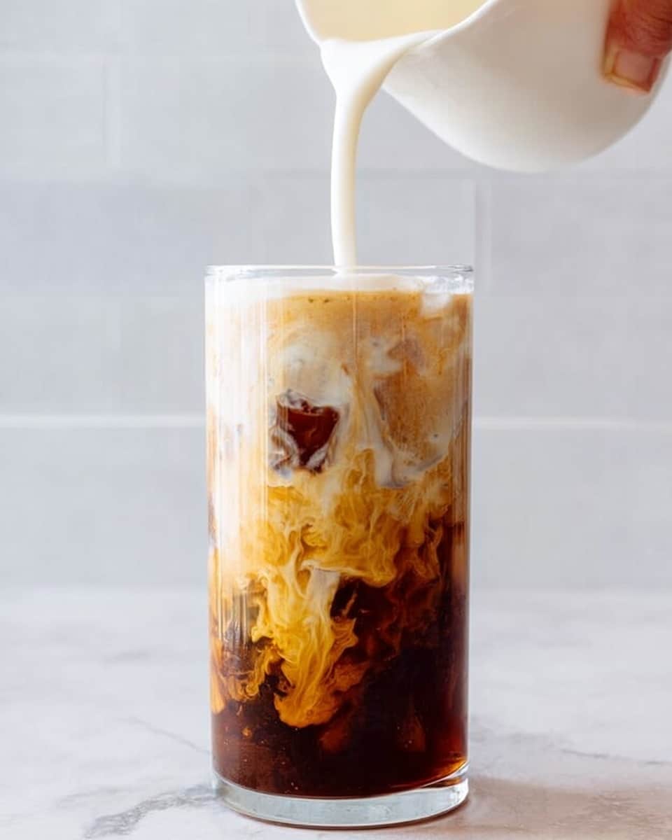 A clear glass filled with three visible layers: the bottom layer is dark brown iced coffee with ice cubes, the middle layer shows swirling light caramel brown cream mixing into the coffee, and the top layer is a thick, creamy white foam that sits smooth at the surface. The glass is placed on a white marbled surface with a soft, light background. The cream swirling creates a marbled, flowing texture inside the glass, contrasting with the solid dark coffee below. Photo taken with an iphone --ar 4:5 --v 7
