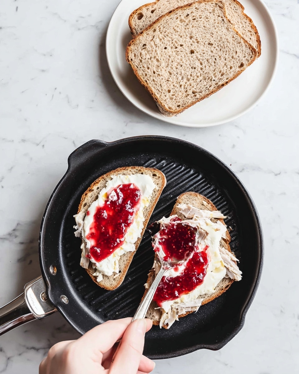 Two slices of brown bread with visible air holes are placed inside a black grill pan on a white marbled surface. Each slice has a layer of white soft cheese spread unevenly, creating a thick, creamy texture. On top of the cheese, there is a bright red jam layer, glossy and slightly spread, with some areas thicker than others. A woman's hand is holding a spoon, adding more jam to one slice, with the spoon tilted and resting on the cheese. Above the pan, on a clean white plate, two thicker slices of the same brown bread stand alone without toppings. The whole setting is lit with natural light, highlighting the textures of the bread, cheese, and jam. Photo taken with an iphone --ar 4:5 --v 7