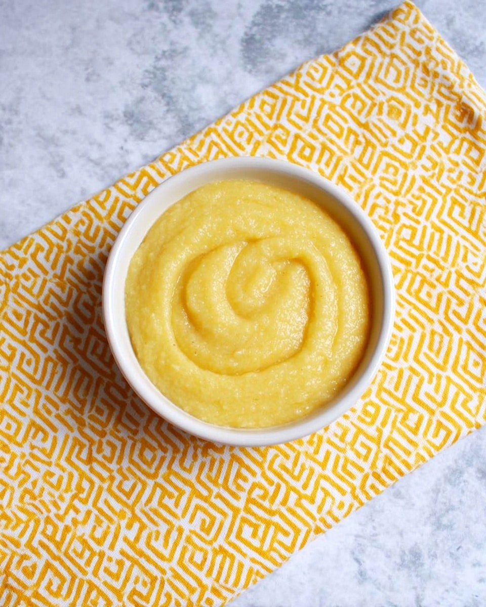 A small white bowl filled with smooth, thick yellow cornmeal mush swirled in a circular pattern on top, placed on a folded yellow napkin with a white geometric pattern, all set on a white marbled surface photo taken with an iphone --ar 4:5 --v 7