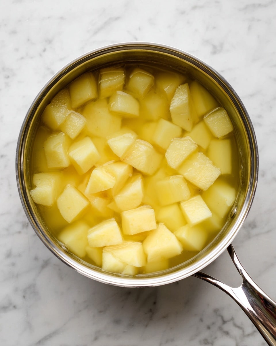 A top view of a silver pot filled with clear yellowish water and many large, light yellow potato chunks soaking inside. The pot is on a white marbled surface, and its shiny silver handle stretches out to the right side of the image. The potato pieces vary in shape but are mostly cubed, surrounded by the calm water inside the pot. Photo taken with an iphone --ar 4:5 --v 7
