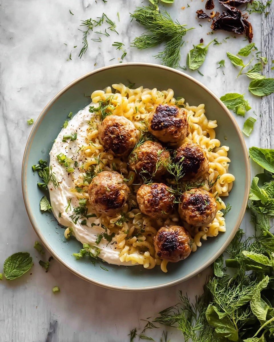 A white bowl with four golden brown meatballs placed on the right side, some fresh green leaves scattered over and around them. On the bottom left is a thick, creamy white sauce with a few dark red sun-dried tomato pieces on top, and a silver spoon rests in the sauce holding one meatball. Behind the meatballs is a layer of yellowish pasta mixed with herbs and a few green olives visible. The bowl sits on a white marbled surface with a few fresh mint leaves scattered nearby. Photo taken with an iphone --ar 4:5 --v 7