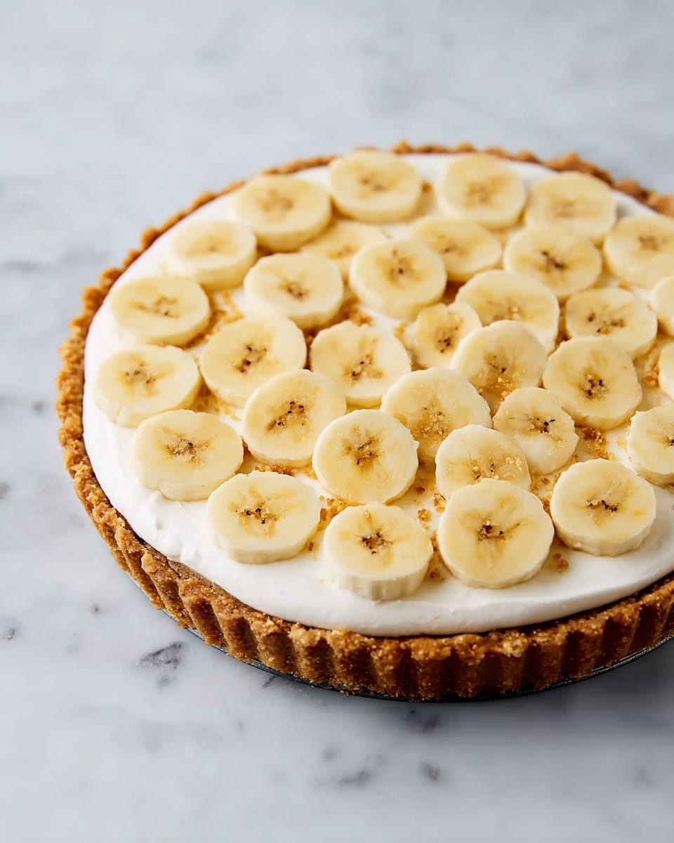 A round tart with a thick golden brown crust forms the base layer. On top of the crust, there is a smooth, thick layer of white cream spread evenly. Over the cream, there are many round banana slices scattered in a loose, random pattern, showcasing their soft yellow color with small brown spots. The tart sits on a flat white marbled surface. photo taken with an iphone --ar 4:5 --v 7