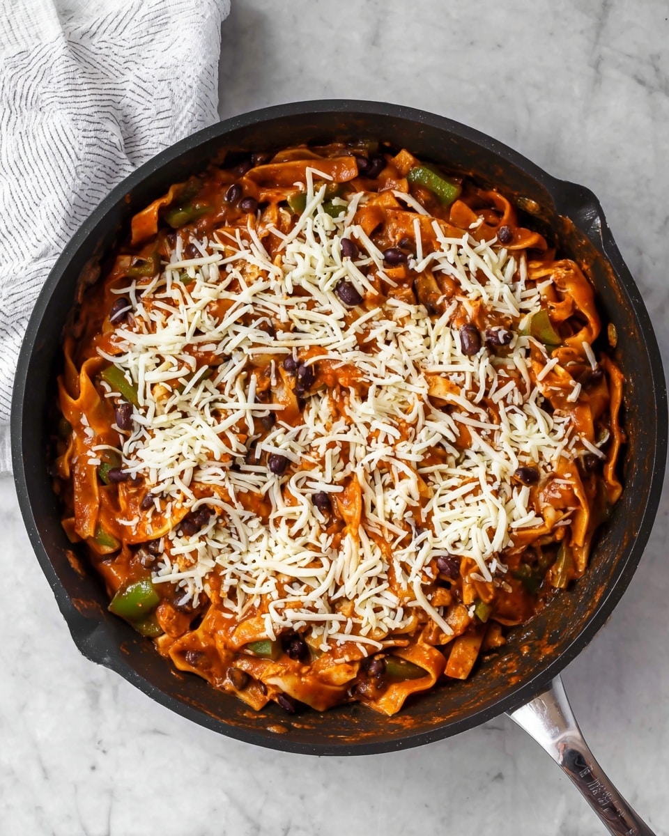 The image shows a black pan filled with cooked pasta mixed with black beans and green bell pepper pieces, all covered in a thick reddish sauce. On top, there is a layer of shredded white cheese scattered over the pasta. The pan is placed on a white marbled surface with a white striped cloth partly visible on the top left side. This dish looks warm and hearty with a mix of soft and slightly chunky textures. Photo taken with an iphone --ar 4:5 --v 7