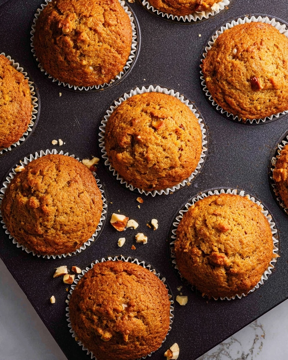 The image shows a close-up top view of nine baked muffins arranged in a dark non-stick muffin tray. Each muffin has a warm golden-brown color with a slightly textured and cracked surface, showing tiny pieces of nuts or fruits inside. The muffins are in white paper liners that create a ruffled edge around each muffin's base, contrasting with the dark tray. Small crumbs and bits of nuts are scattered on the tray between the muffins. The background is a white marbled texture. photo taken with an iphone --ar 4:5 --v 7