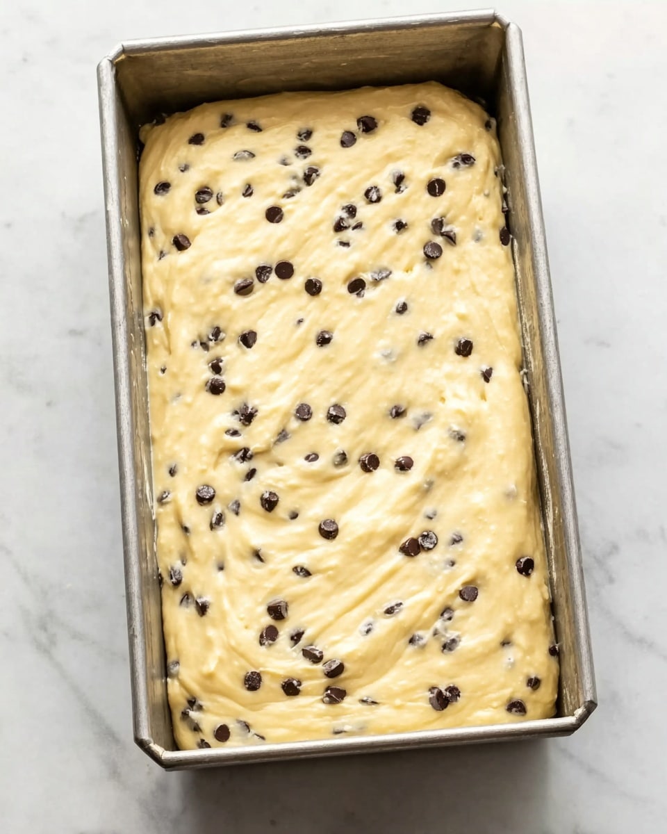 A metal rectangular pan filled with smooth, creamy batter spread evenly across its surface, dotted with small dark chocolate chips scattered throughout. The batter has a light yellow color with a slightly glossy texture. The pan is placed on a white marbled surface, creating a clean and fresh background. photo taken with an iphone --ar 4:5 --v 7