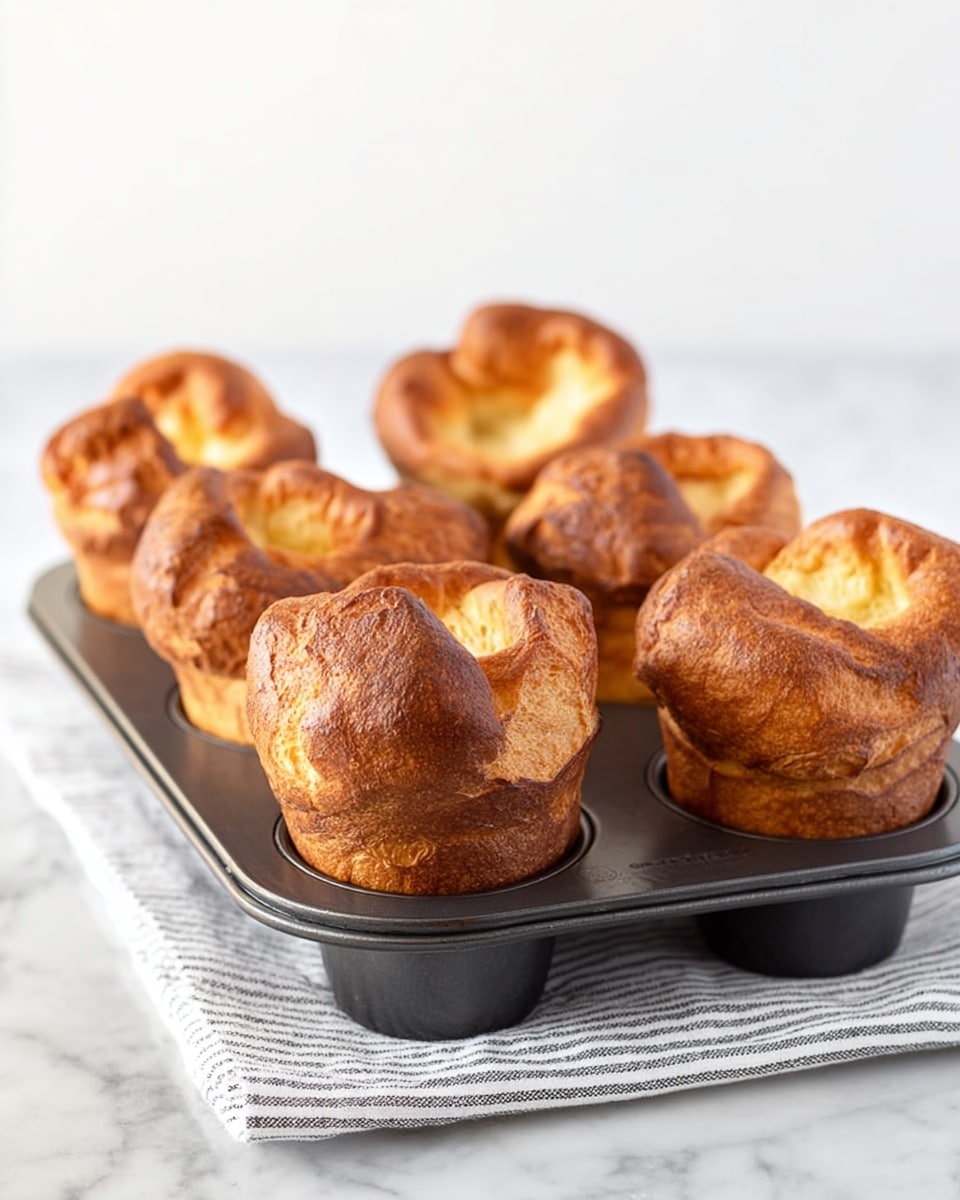 The image shows six golden-brown popovers rising above a black metal popover pan with six rounded cups. Each popover has a puffy, irregular shape with crisp, slightly darker edges and a lighter, soft inside visible at the base. The pan is placed on a white marbled surface with a folded striped cloth underneath it. The background is a clean white, making the warm tones of the popovers stand out. photo taken with an iphone --ar 4:5 --v 7