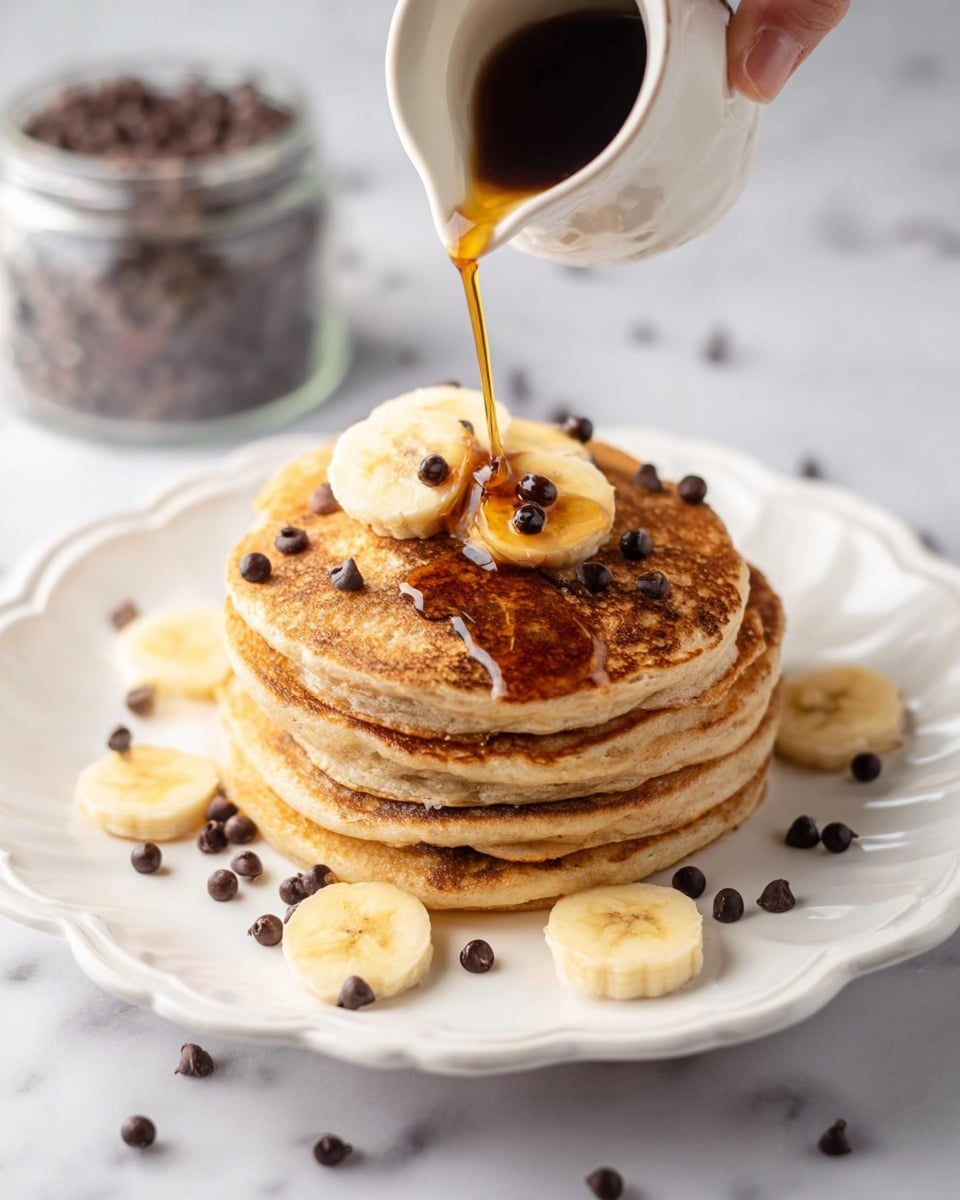 A stack of five golden-brown pancakes sits on a white scalloped plate placed on a white marbled surface. The pancakes have a soft, fluffy texture with visible tiny air holes. The top pancake is decorated with round banana slices and small chocolate chips scattered on them. Maple syrup gently glistens over the pancakes and pools slightly at the base, covering more banana slices. A fork is lifting three small stacked pieces of pancake near the plate's right edge, showing the airy inside. photo taken with an iphone --ar 4:5 --v 7