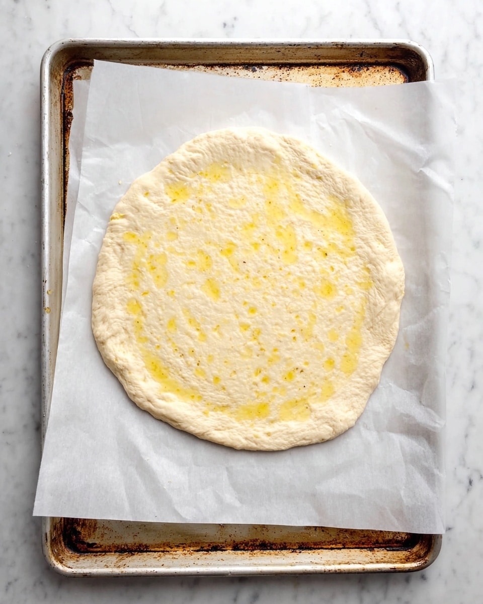 A round raw pizza dough with light beige color and slightly uneven edges lies flat on top of white parchment paper on a metal baking sheet. The dough is a single thick layer with a slightly rough texture and has small spots of yellow oil sprinkled across its surface, mainly in the middle. The baking sheet shows signs of use with rust and stains around the edges, placed on a white marbled surface. photo taken with an iphone --ar 4:5 --v 7