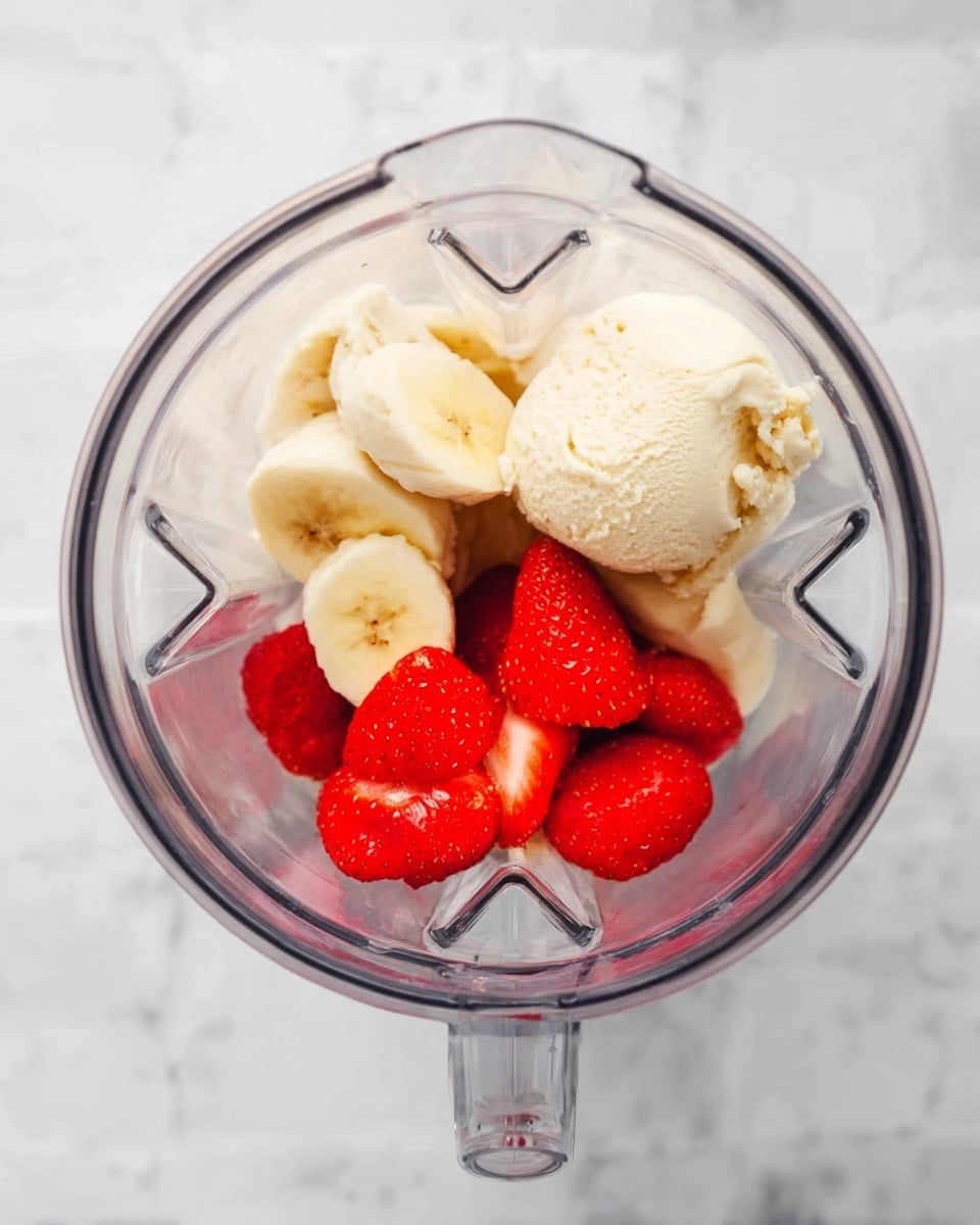 A clear blender jar is seen from above, filled with three main layers of ingredients. At the bottom, there are three bright red strawberries with a smooth texture, placed side by side. Above the strawberries, a thicker layer of pale yellow banana slices is stacked, showing the soft and slightly moist texture. On top of the bananas, there are two large scoops of a creamy, light beige substance, possibly ice cream, with a soft and slightly rough texture. The blender is set on a white marbled surface. photo taken with an iphone --ar 4:5 --v 7