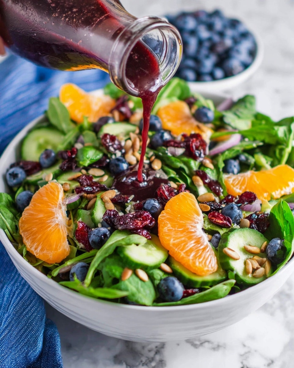 A white bowl filled with a colorful salad sits against a white marbled surface. The salad has a mix of green leafy spinach and lettuce at the base, layered with bright orange mandarin orange slices, dark blue blueberries, slices of green cucumber, and scattered dried red cranberries. Light brown sunflower seeds are sprinkled on top. A woman's hand is pouring a thick dark red dressing over the salad from a clear glass bottle, adding a glossy texture to the fruit and greens. Photo taken with an iphone --ar 4:5 --v 7