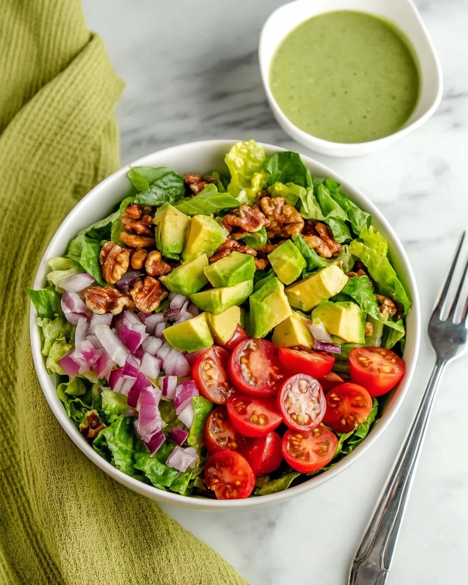 A white bowl filled with a fresh salad showing three main layers: the bottom layer has green leafy lettuce, the middle layer has bright red cherry tomato halves and small pieces of purple onion, and the top layer features chunky yellow-green avocado pieces and scattered golden brown walnut pieces. Next to the bowl, there is a white small bowl with green dressing, placed on a white marbled surface along with a silver fork on the right side. A green cloth is partly visible on the left side. Photo taken with an iphone --ar 4:5 --v 7