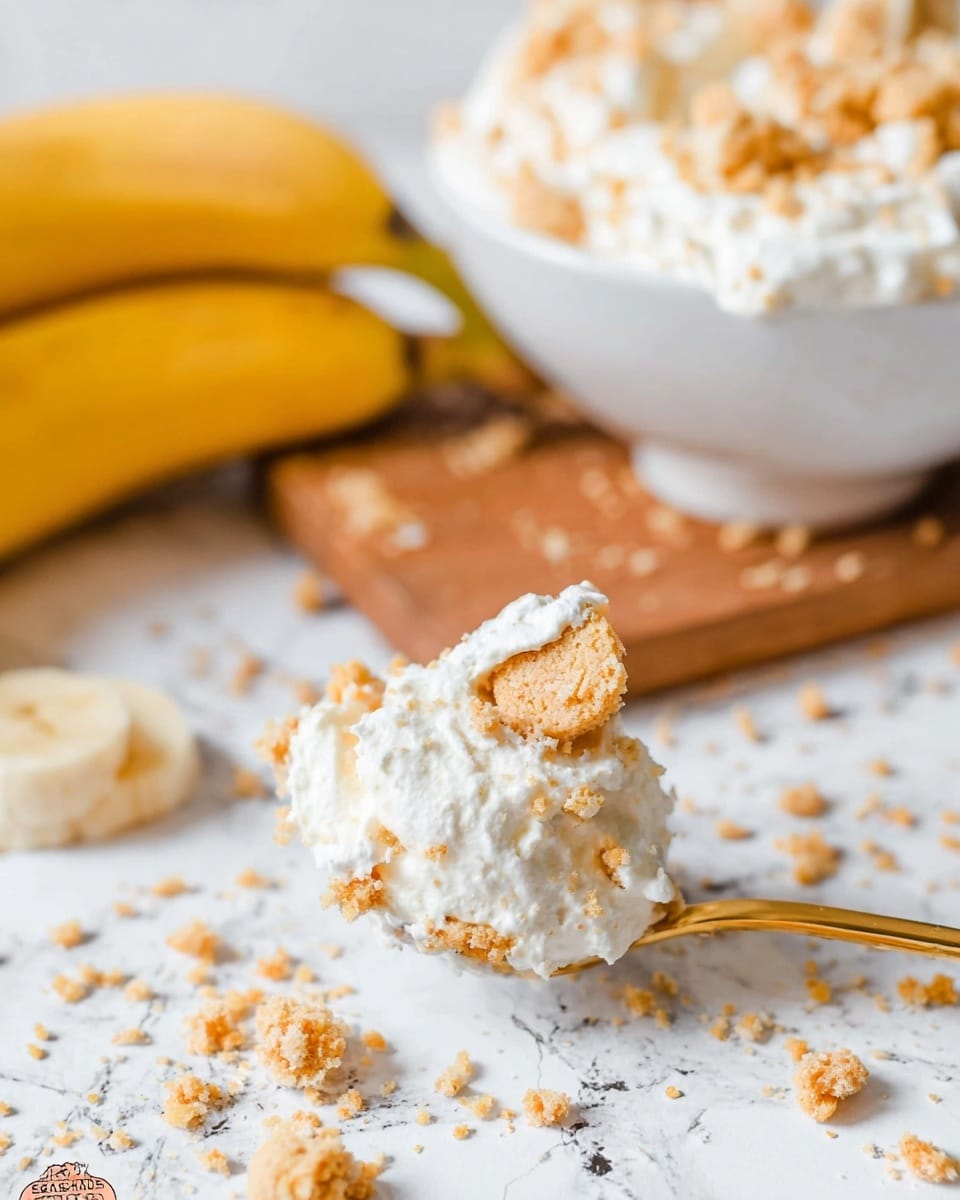 A close-up image shows a small gold spoon holding a creamy, white mixture with visible crumbled light brown cookie pieces. The spoon is placed on a white marbled surface that has scattered cookie crumbs around it. Behind the spoon, there are two yellow bananas lying horizontally on a wooden board, partially blurred. In the background, a white bowl filled with the same creamy mixture and cookie crumbs is visible, resting on the wooden board. The photo taken with an iphone --ar 4:5 --v 7