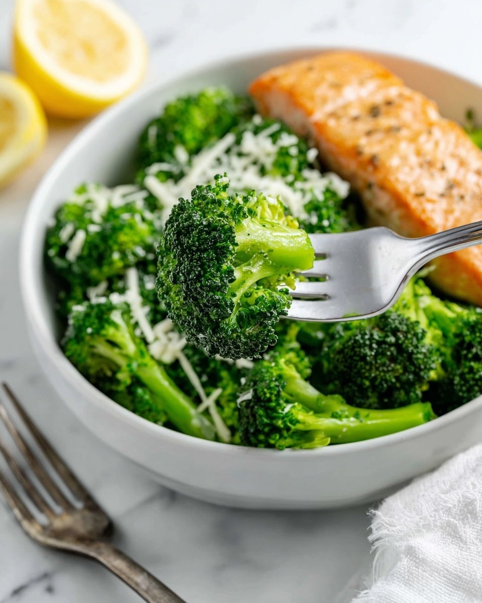 A white round plate sits on a white marbled surface. The plate has two main layers: on the left, a large pile of bright green steamed broccoli florets with a light topping of finely grated white cheese. On the right, a golden brown grilled salmon fillet with a slightly crispy and peppered surface, its edges darkened a little. In the background, there are two silver forks, a yellow wedge of cheese, and two whole garlic bulbs, all placed on the white marbled surface with a soft white cloth loosely folded nearby. Photo taken with an iphone --ar 4:5 --v 7