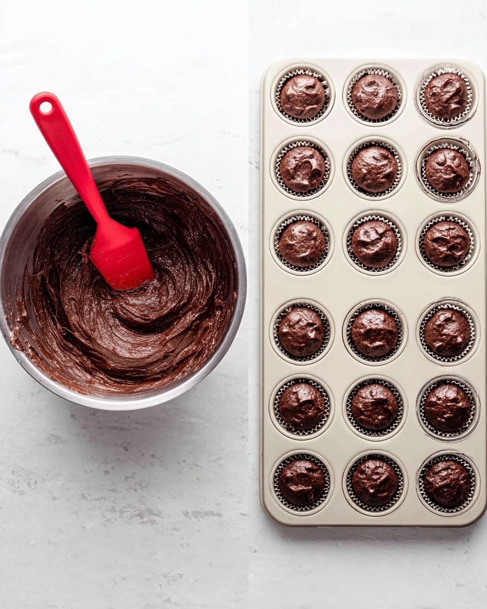 The image shows two items on a white marbled surface side by side. On the left is a metal mixing bowl filled with thick, dark brown chocolate batter with a smooth but slightly uneven texture. A red spatula rests inside the bowl, partially coated with the batter. On the right is a white metal mini muffin pan with 24 small round compartments neatly filled with dollops of the same chocolate batter, each portion roughly equal in size and with textured tops. The surface and arrangement highlight the preparation stage of chocolate mini muffins or cupcakes. Photo taken with an iphone --ar 4:5 --v 7