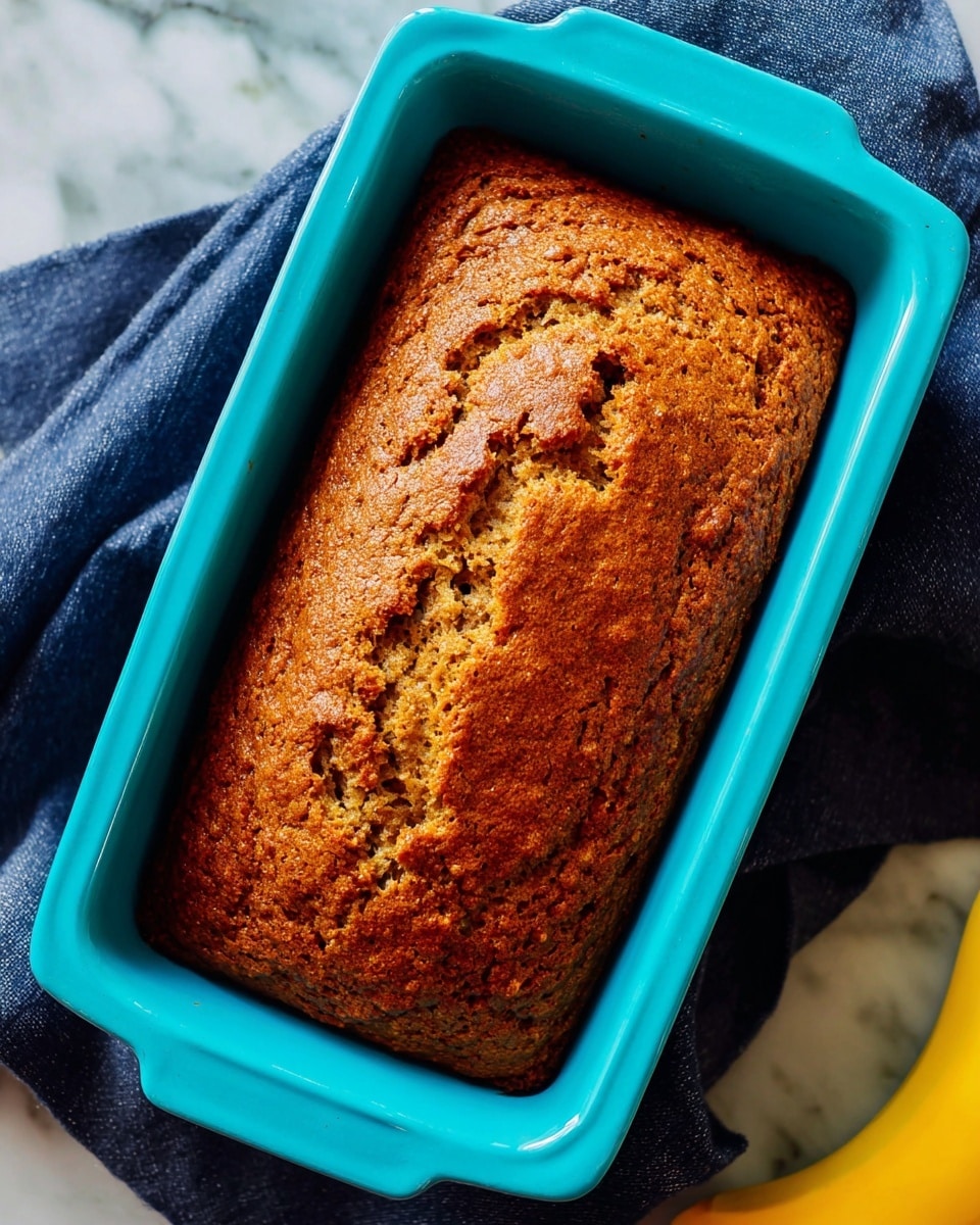 A single loaf of golden brown bread with a textured, slightly cracked top sits inside a bright turquoise rectangular baking pan. The bread has a rich, warm color with darker browned spots scattered on the surface, indicating it is well baked. The pan rests on a dark blue cloth that covers part of a white marbled surface, with a small portion of a ripe yellow banana visible at the bottom right corner of the image. photo taken with an iphone --ar 4:5 --v 7