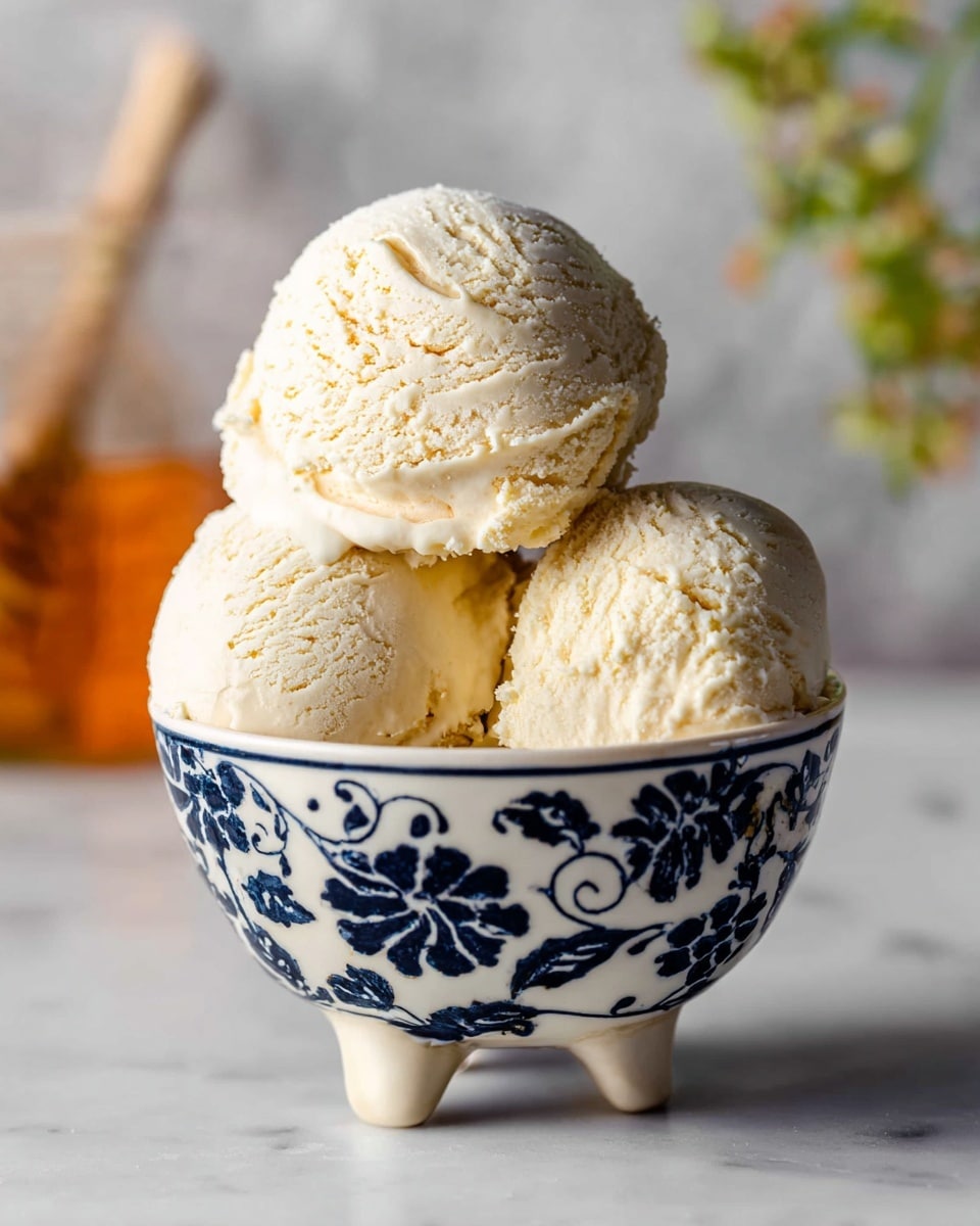 The image shows three scoops of light cream-colored ice cream with a slightly grainy texture, placed in a white cup decorated with dark blue swirling floral patterns and standing on three small legs. The ice cream scoops are stacked neatly inside the cup, with one scoop slightly raised above the others, showing soft and smooth edges. The background features a blurred white marbled surface, with out-of-focus elements in warm amber and green tones adding depth. photo taken with an iphone --ar 4:5 --v 7