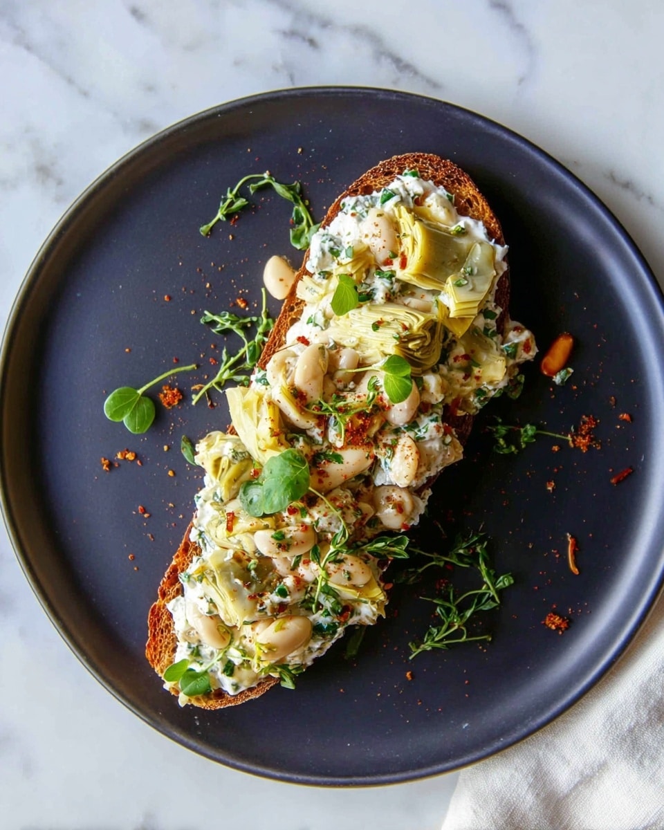 A single long slice of toasted brown bread forms the base, topped with a thick layer of creamy white cheese mixed with light green artichoke hearts and whole white beans. Small bright green herb leaves are scattered on top, along with a light sprinkle of crushed red spice that adds hints of red and orange. The dish is served on a simple white plate with a slight gloss, placed on a white marbled surface with a soft white marbled texture visible under the plate. Photo taken with an iphone --ar 4:5 --v 7