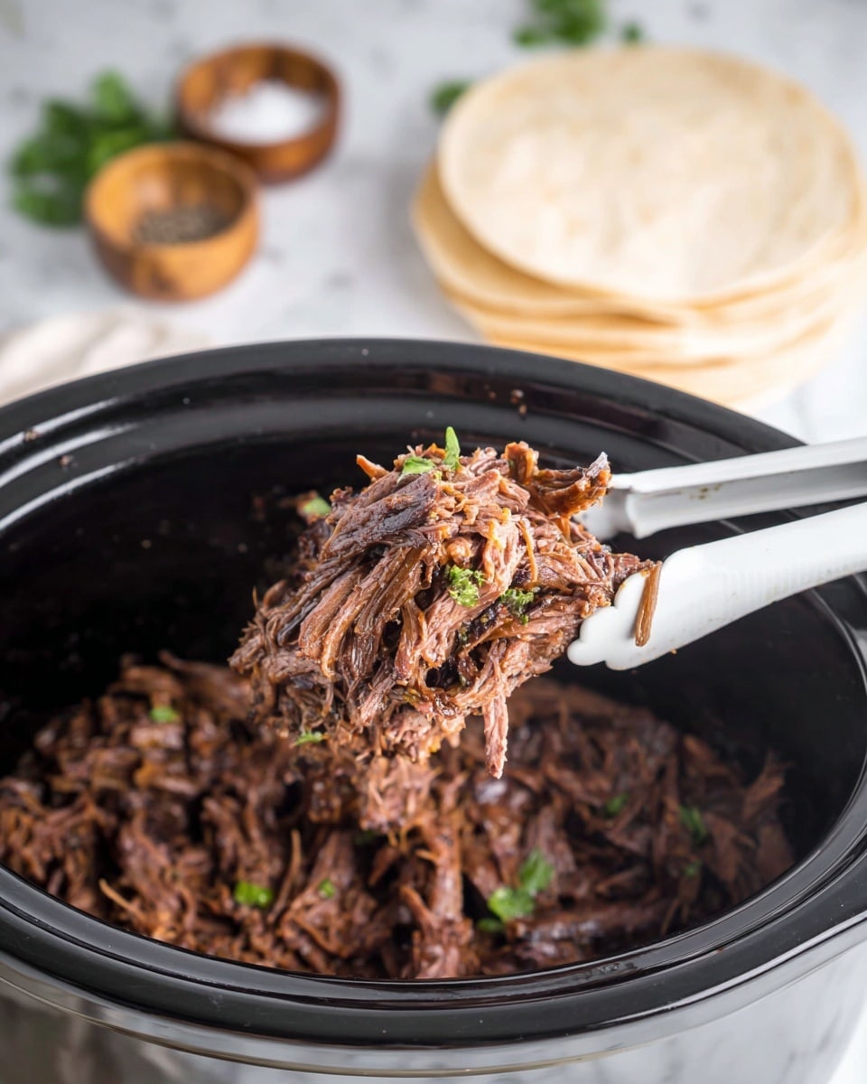 The image shows a close-up of shredded slow-cooked beef held by white tongs above a black slow cooker filled with more shredded beef. The beef is dark brown with a tender texture and bits of green herbs mixed throughout. In the background, there is a stack of round white corn tortillas on a white marbled surface along with small wooden bowls of salt and pepper, giving a simple, rustic feel. The colors contrast between the dark meat and the white surroundings, highlighting the rich texture of the beef. photo taken with an iphone --ar 4:5 --v 7
