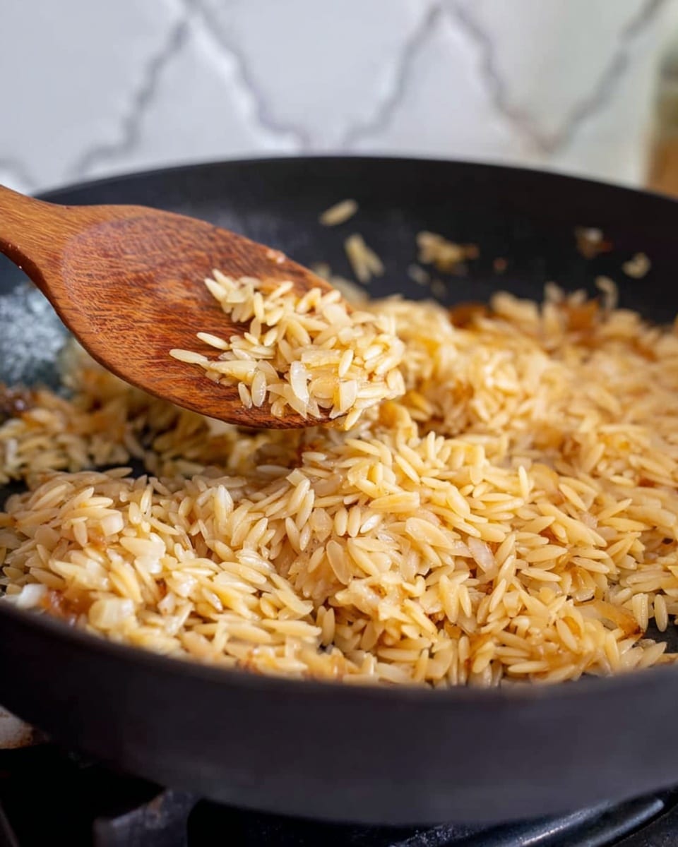 A close-up view of a black pan on a white marbled surface filled with golden toasted orzo pasta mixed with small pieces of cooked white onion. A wooden spoon is stirring the orzo, lifting some of the mixture, showing the toasted texture and light brown color of the orzo along with translucent onion bits. The background has a blurred white backsplash with a subtle pattern. Photo taken with an iphone --ar 4:5 --v 7