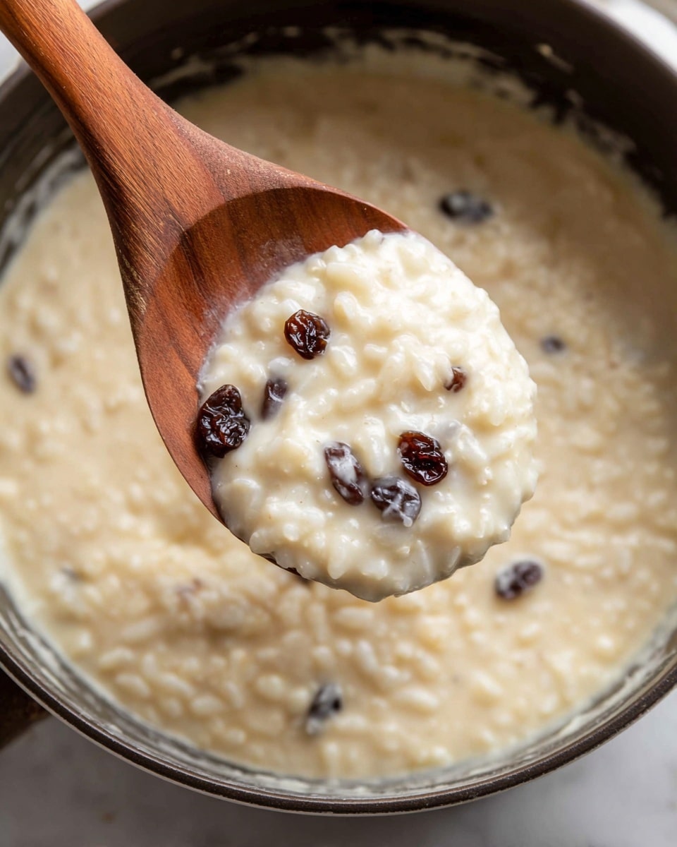 A close-up image of a creamy rice pudding with a soft texture, showing small, plump white rice grains mixed with dark brown raisins. The pudding is thick and smooth with a pale cream color, and it is held in a wooden spoon above a pot filled with the same mixture. The pot is dark on the outside with smears of the pudding along the inner edge. The scene is set on a white marbled surface. photo taken with an iphone --ar 4:5 --v 7